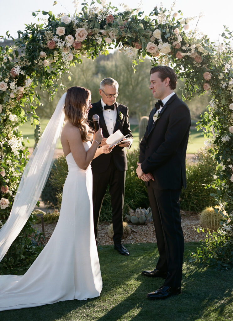 Bride speaking into microphone across from groom at outdoor wedding ceremony, both in front of officiant and curved floral and greenery arch.
