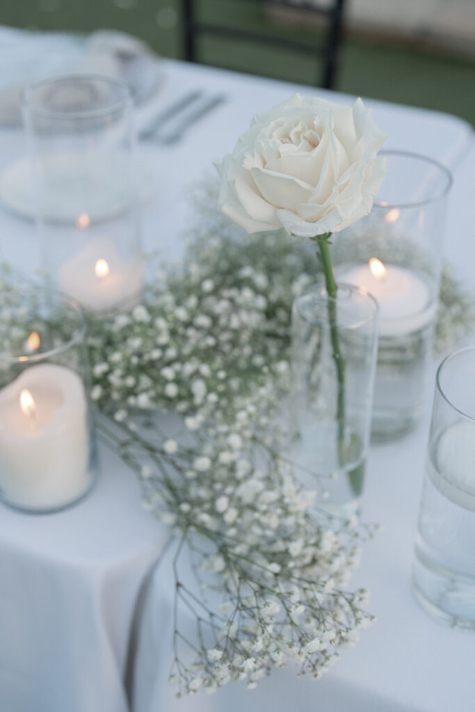 White rose in clear glass modern bud vase with baby's breath and candles.