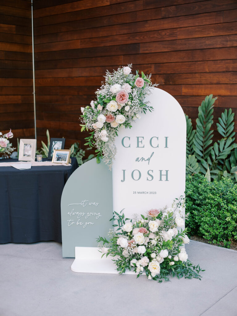 Custom wedding welcome sign with floral arrangements installed on the top and on the ground in front of sign of white and blush floral.