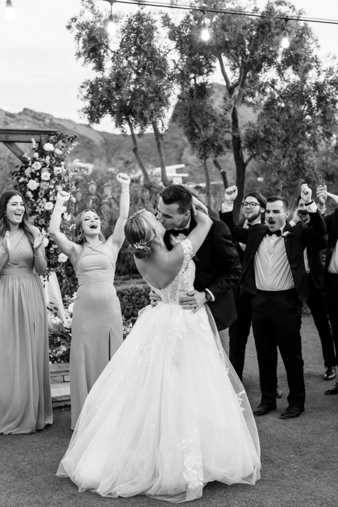 Bride and groom kissing with wedding party celebrating behind them.