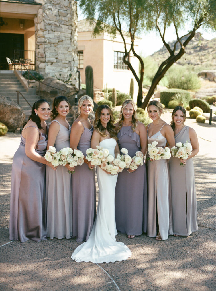 Bride standing in live with bridesmaids wearing taupe dresses, all holding bouquets of white and blush floral.