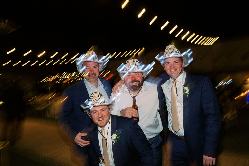 Three men in blue suits at wedding reception with white cowboy hats on.