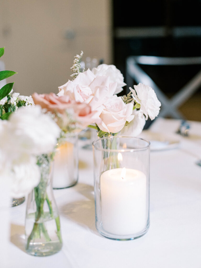 Candles and white and blush floral bud vases on white linen reception table at wedding.