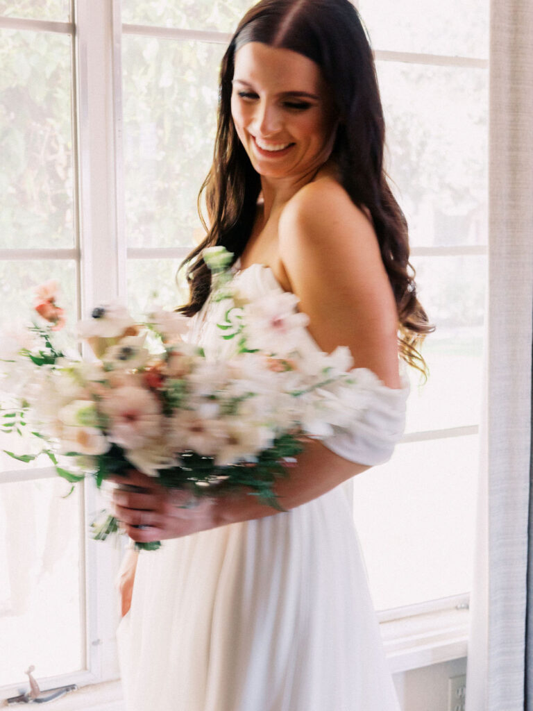 Bride smiling, looking down at bouquet of white and pink standing in front of window during day.