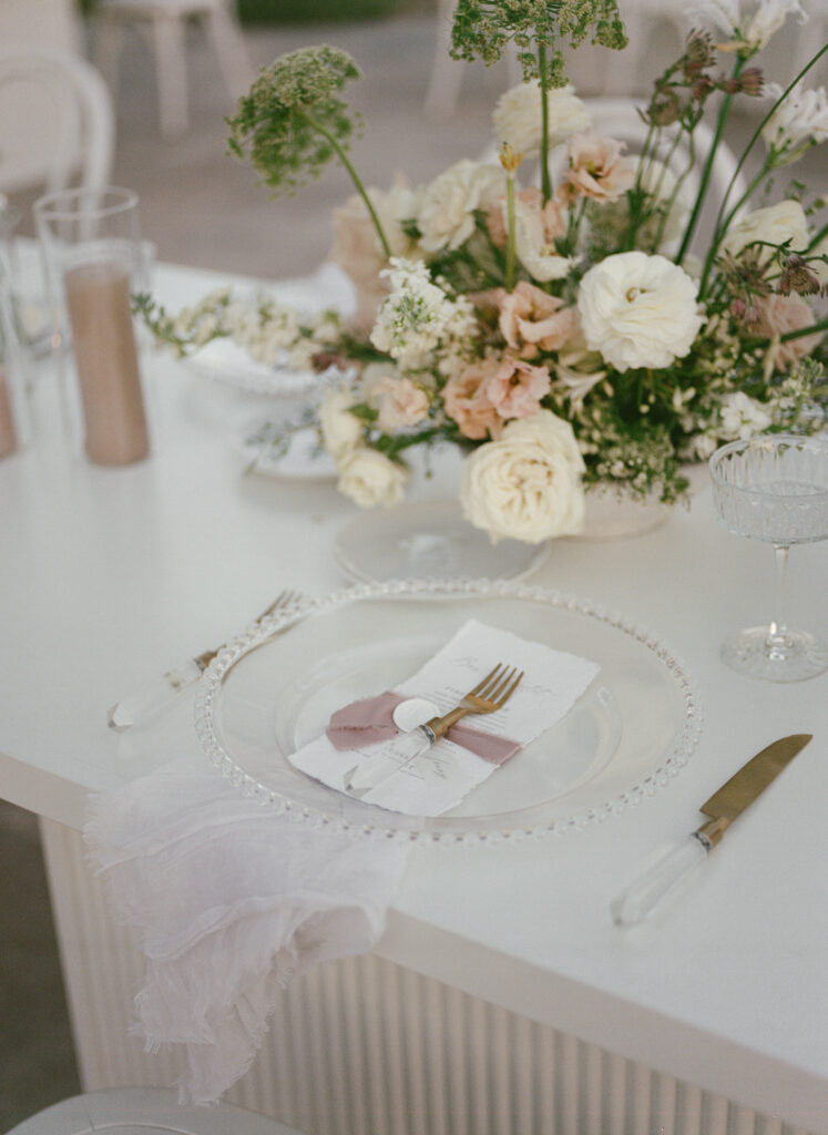 Wedding reception table with glass glass and plate and unique centerpieces of white and blush floral.