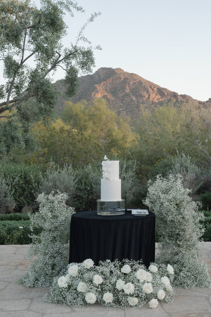 White tiered wedding cake on round table with black linens surrounded by baby's breath and white roses.