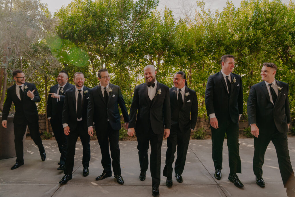 Groom walking with groomsmen in black suits outside with trees behind them.