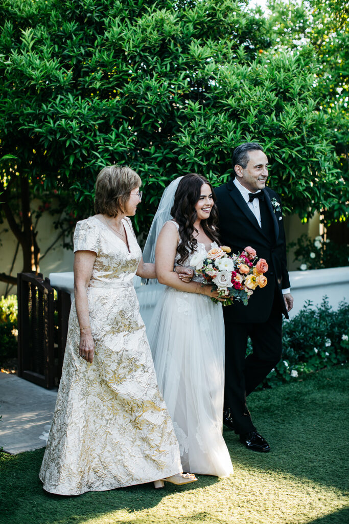 Bride entering outdoor wedding ceremony aisle with man and woman on either side of her and holding bouquet.