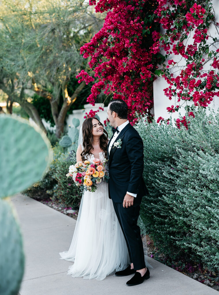 Bride holding bouquet smiling up at groom looking at her in black suit both standing on sidewalk in lush desert landscape.