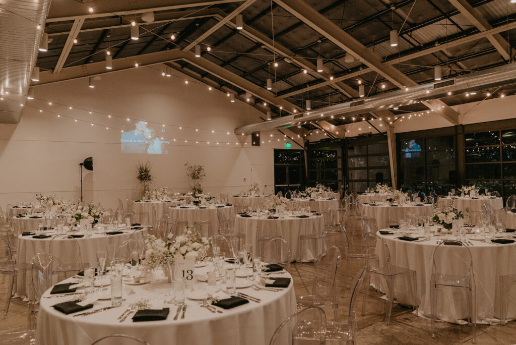 The Clayton House wedding reception space with round tables with white flowers centerpieces, white linens, and clear acrylic chairs.
