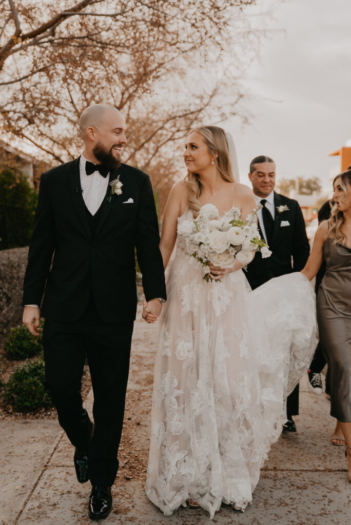 Bride and groom smiling at each other and holding hands walking down sidewalk with groomsmen and bridesmaids behind them.