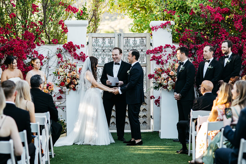 Outdoor wedding ceremony at El Chorro in front of doors with bride and groom standing in front between flower arrangements on acrylic pillars with officiant.