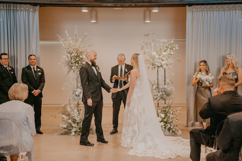 Bride and groom holding hands at wedding ceremony altar space in front of officiant with white flowers pillars framing them on sides.