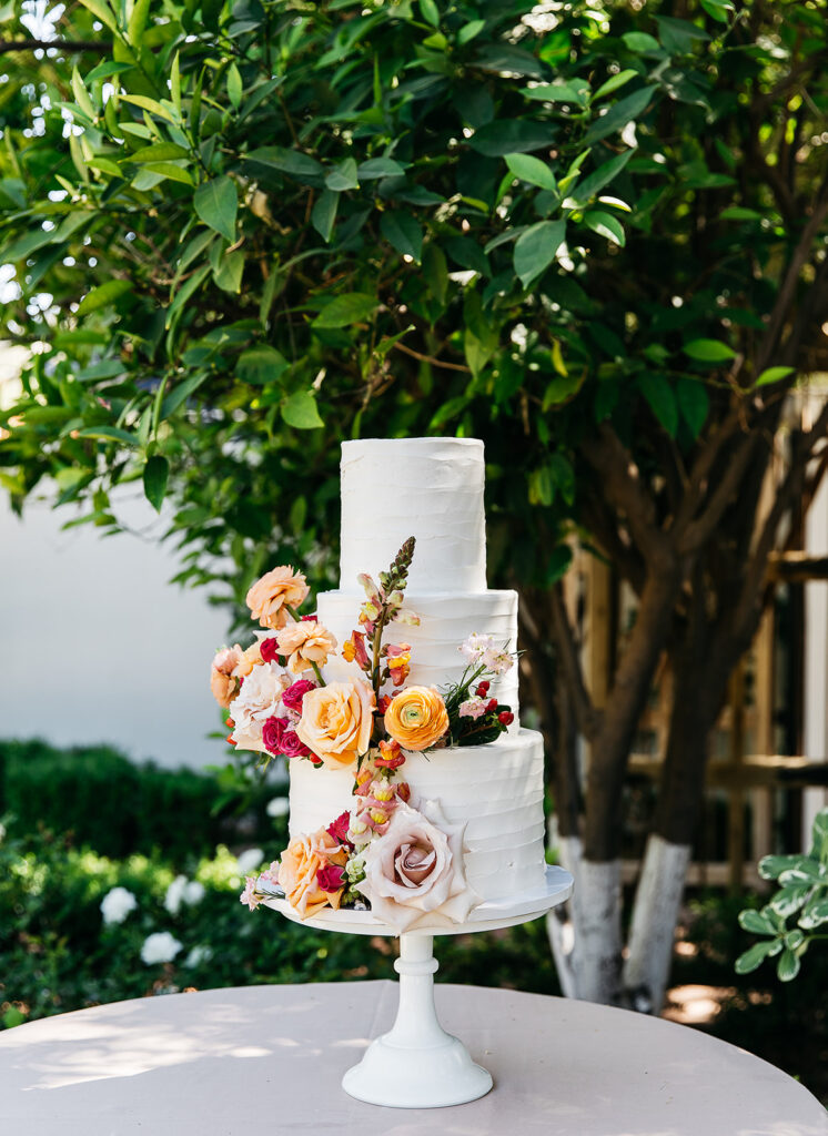 Three tiered white wedding cake with colorful floral added to bottom two tieres on tall cake stand.