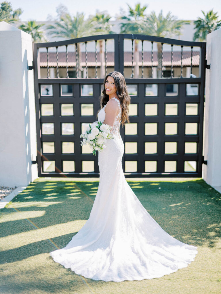 Bride standing in front of metal gate at Scottsdale Resort looking over shoulder at camera holding bouquet of white flowers and greenery.