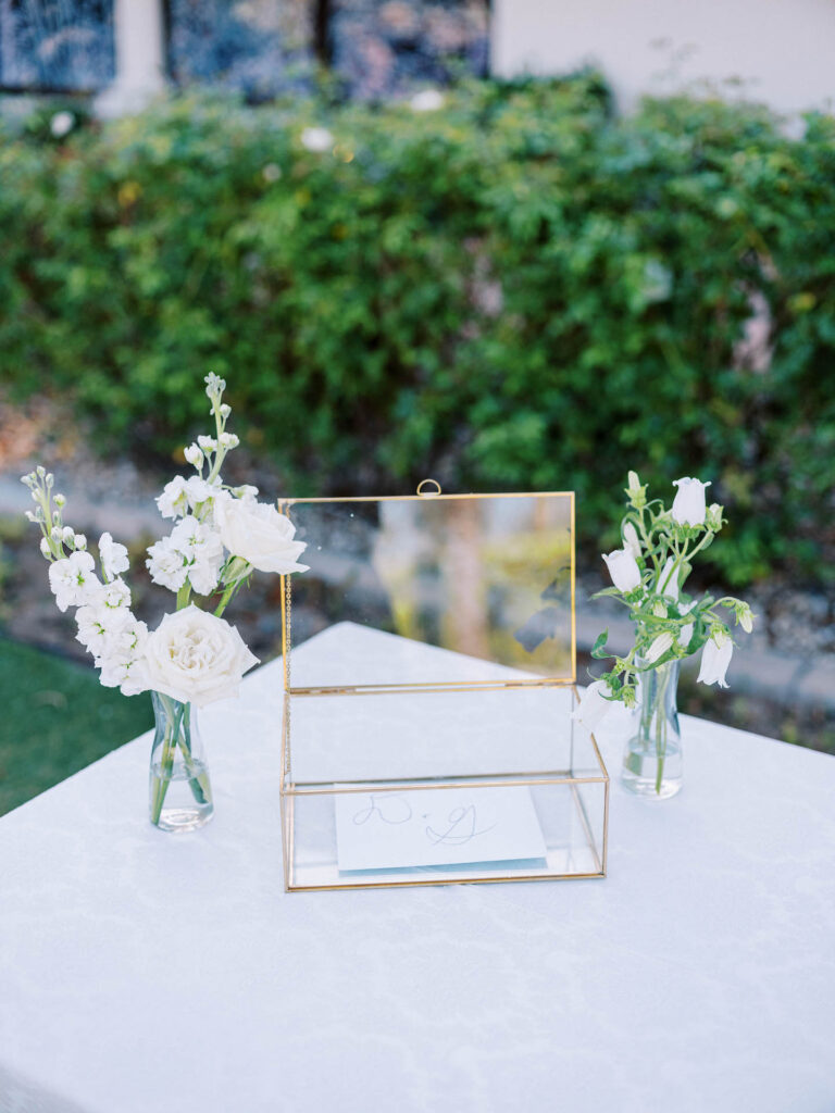 Welcome table glass envelope box with white florals in bud vases on either side.