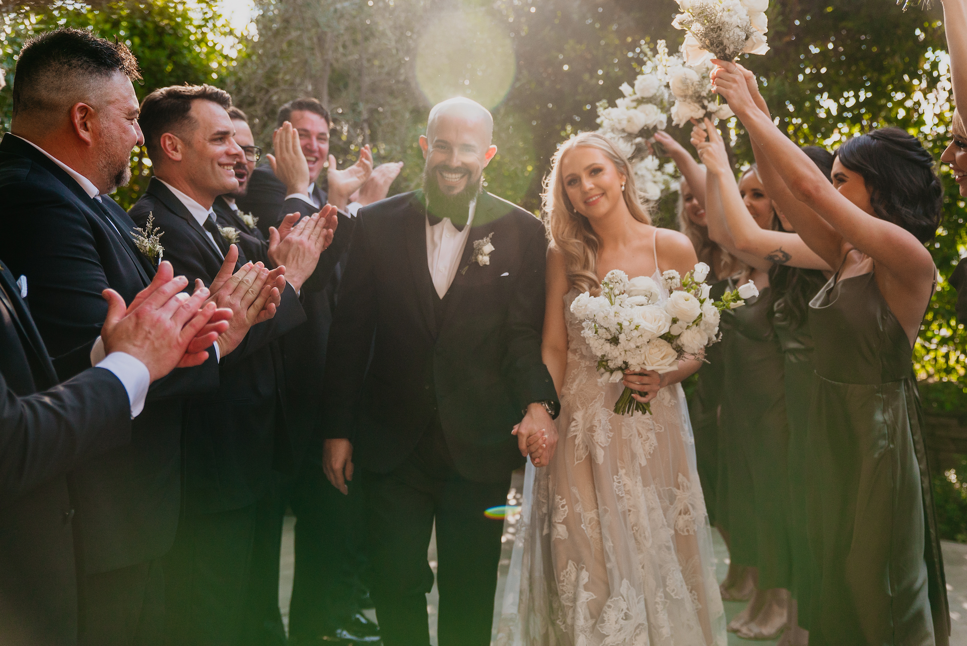 Bride and groom holding hands, smiling, walking between groomsmen and bridesmaids lining the sides and clapping.