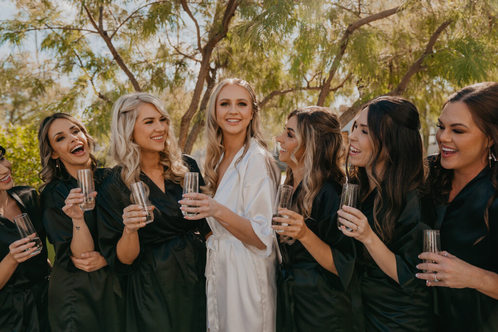 Bride in white bathrobe with bridesmaids in black bathrobes with glasses of champagne standing together outside.