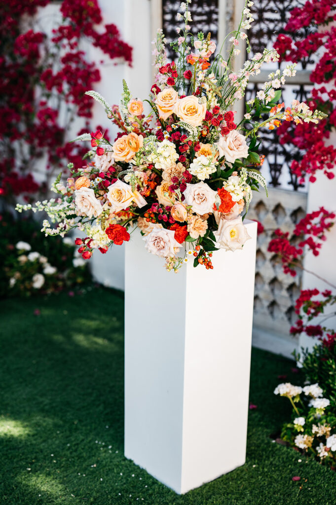 Large reaching floral arrangement on white acrylic pillar at outdoor wedding ceremony.