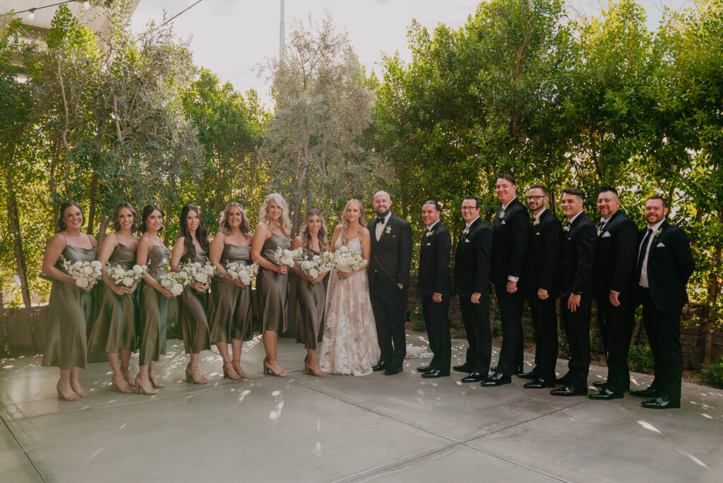 Wedding party of bridesmaids and groomsmen standing on either side of bride and groom in center, standing outside.
