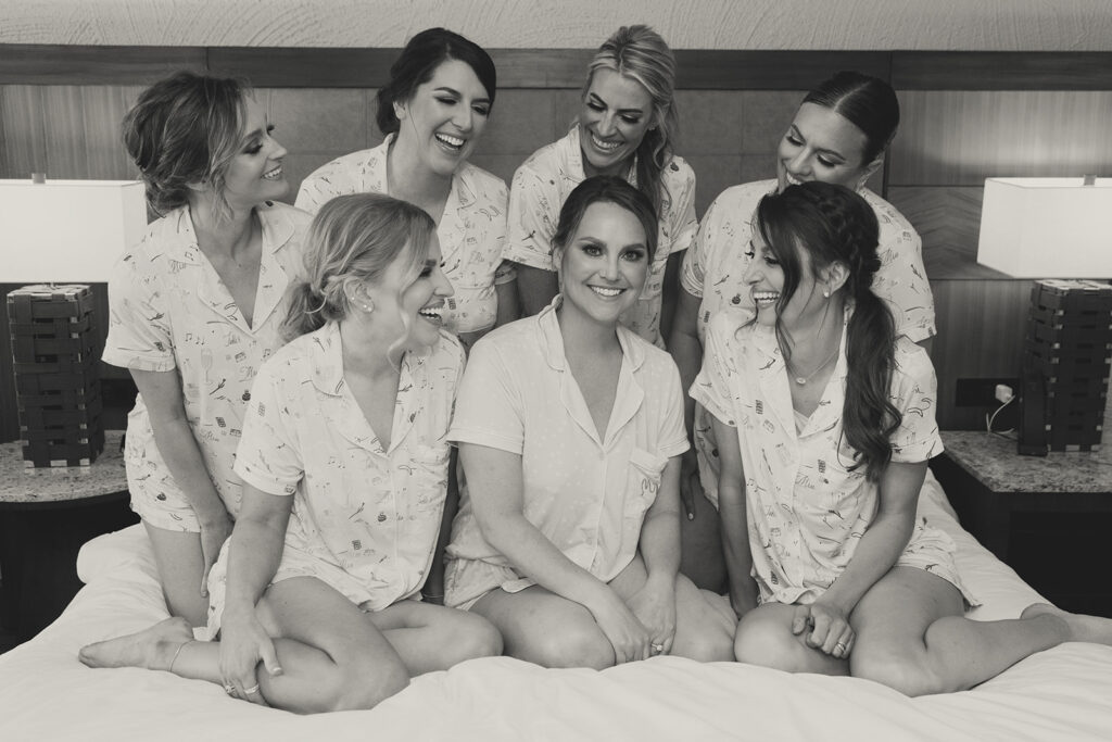 Bride with bridesmaids sitting on a bed, all smiling in pajamas getting ready for wedding.