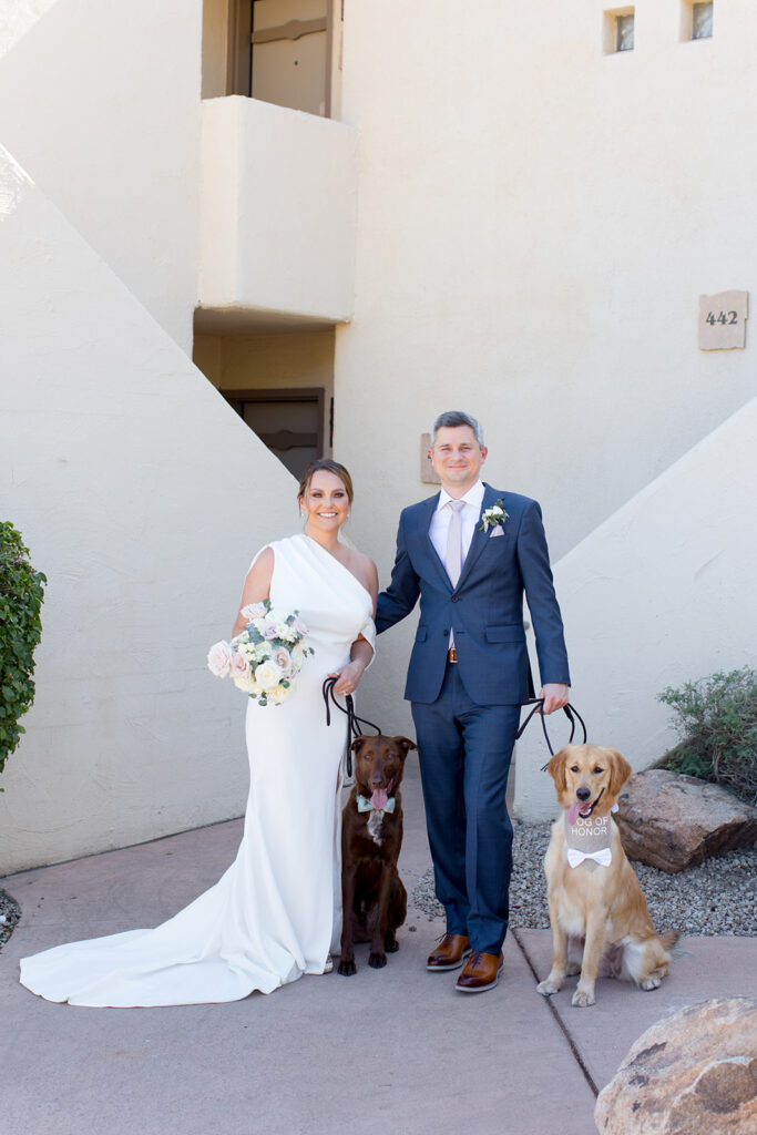 Bride and groom smiling, standing outside white building holding leashes of a dog each.