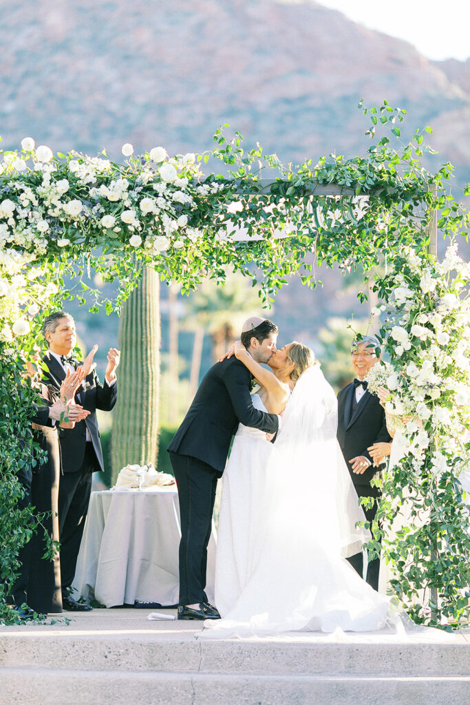 Bride and groom kissing under floral arch at outdoor wedding ceremony.