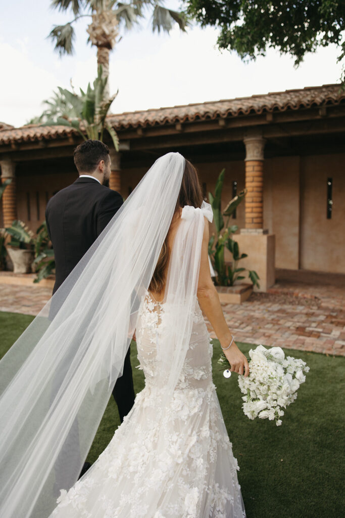 Bride and groom walking away at on Royal Palms lawn, bride with long veil trailing.