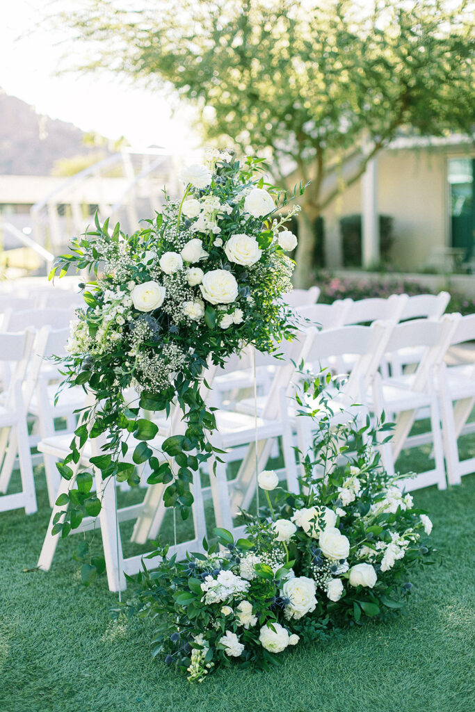 Back of outdoor wedding ceremony floral arrangements of white floral and greenery, some on grass and others on clear acrylic stand.