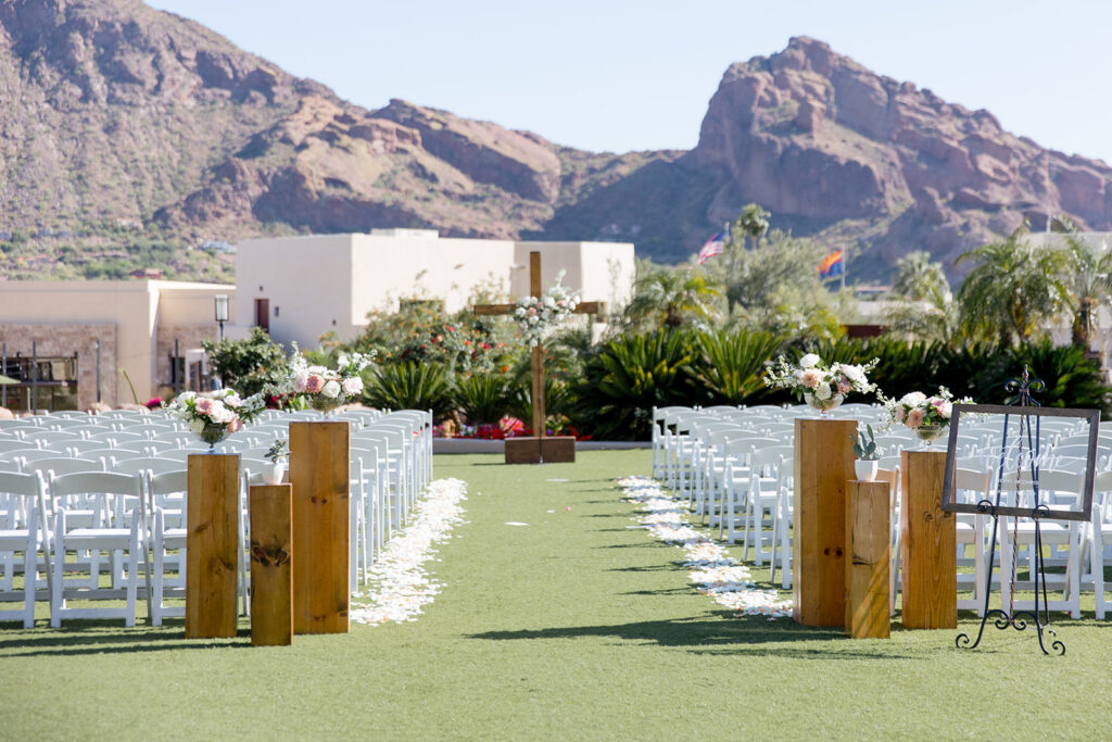 Outdoor wedding ceremony at Camelback Inn with Camelback Mountain in distance, aisle entrance arrangements at back of aisle, white rose petals lining the aisle and wood cross with floral installation at altar space.
