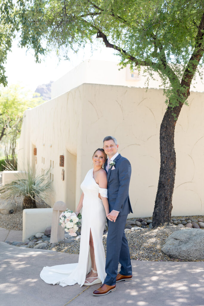 Bride resting with her back against groom holding bouquet, both smiling, standing in front of southwest style architecture building and desert tree.