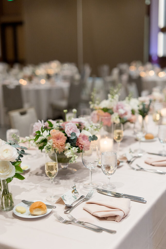 Wedding reception table with glass vessel centerpiece of white and blush roses and bud vases on blush linens.