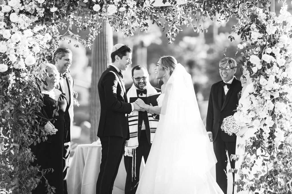 Bride and groom holding hands, smiling at each other, with people around them under floral arch at wedding ceremony.