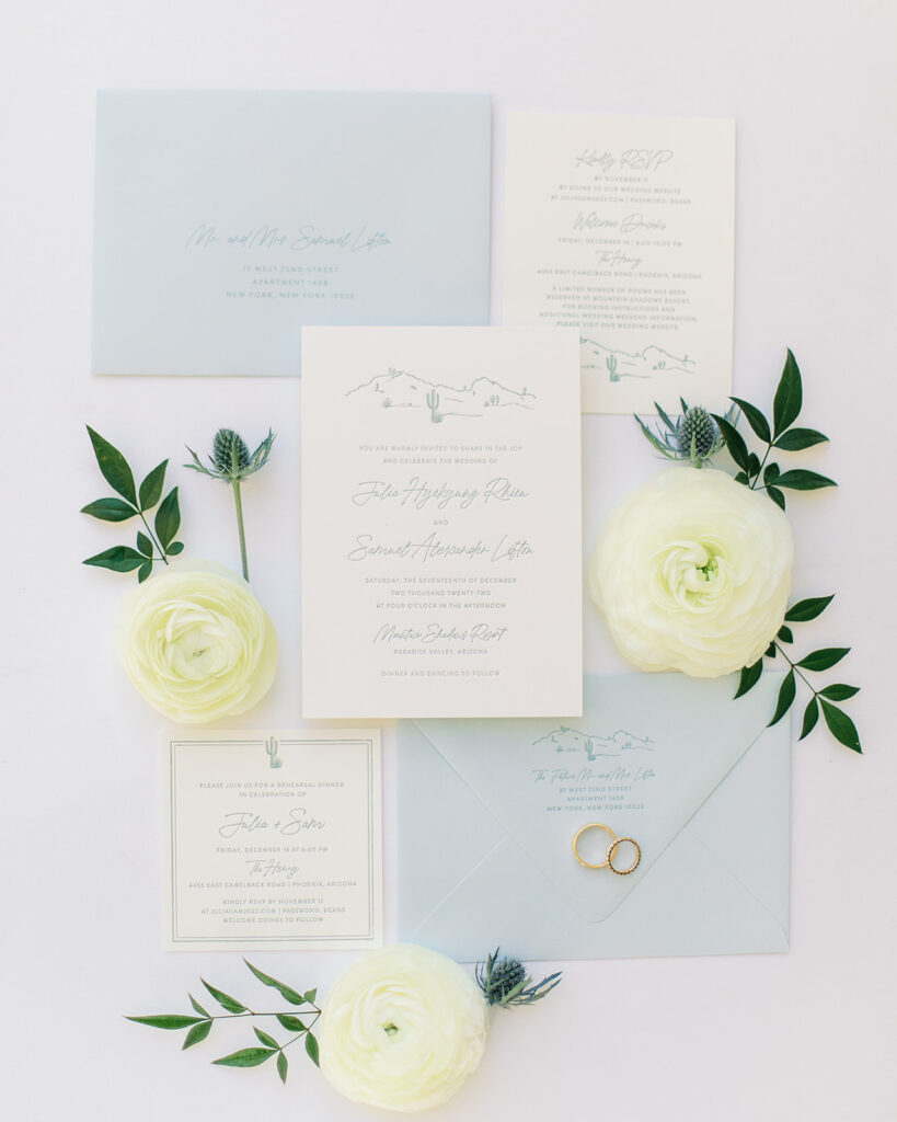 Wedding invitation flat lay of white and soft blue with ranunculus and thistle accents.