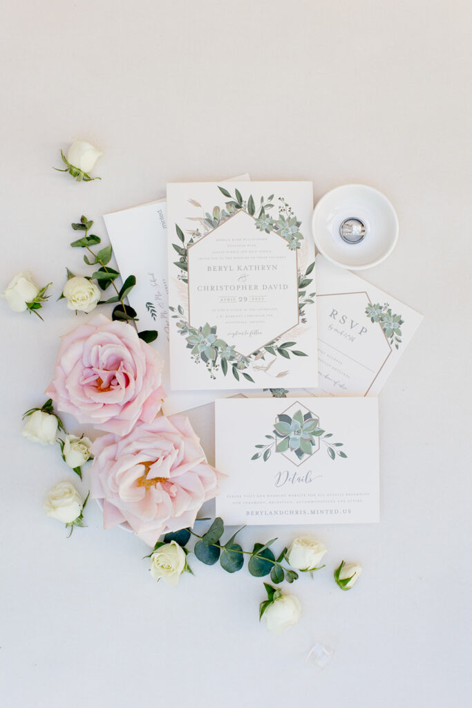 White wedding invitation suite with succulent and greenery details in a flat lay with roses and eucalyptus and weding rings in a bowl.