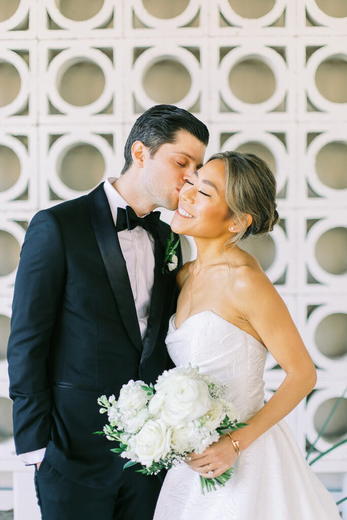 Groom kissing bride on the cheek, both smiling. Bride holding white flowers bouquet.