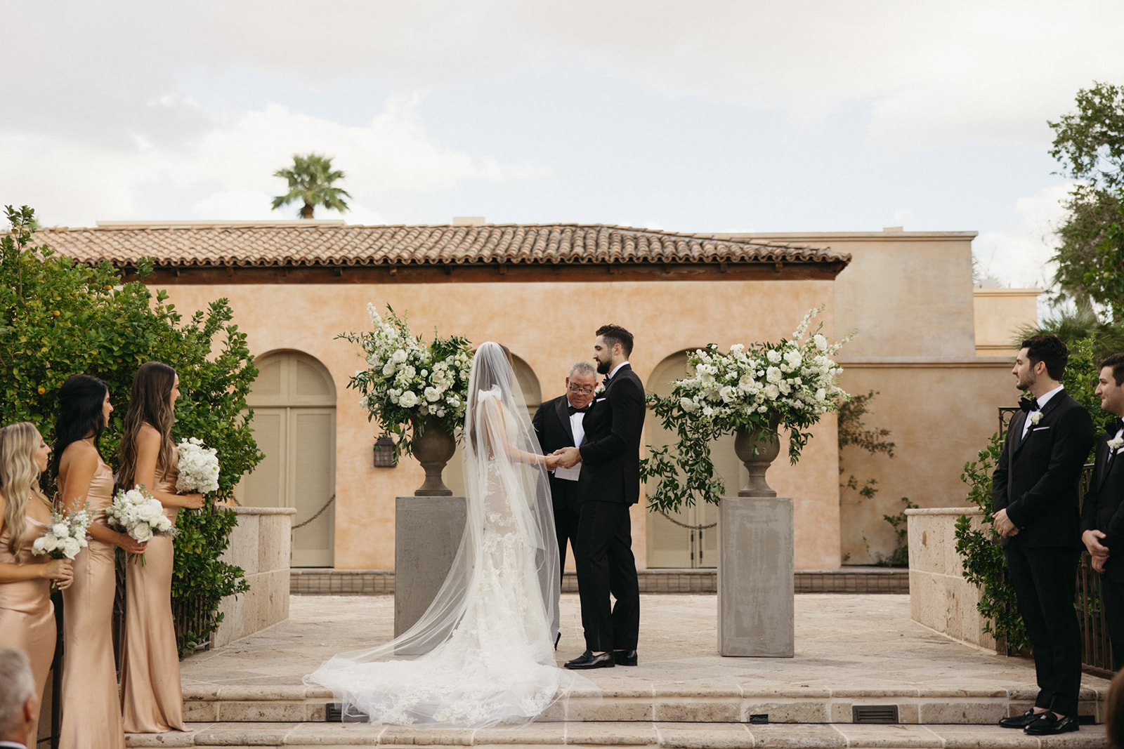 Bride and groom at outdoor wedding ceremony at Royal Palms up on steps between cement pillars with large white floral arrangements framing them with officiant behind them and bridesmaids and groomsmen to their respective sides.