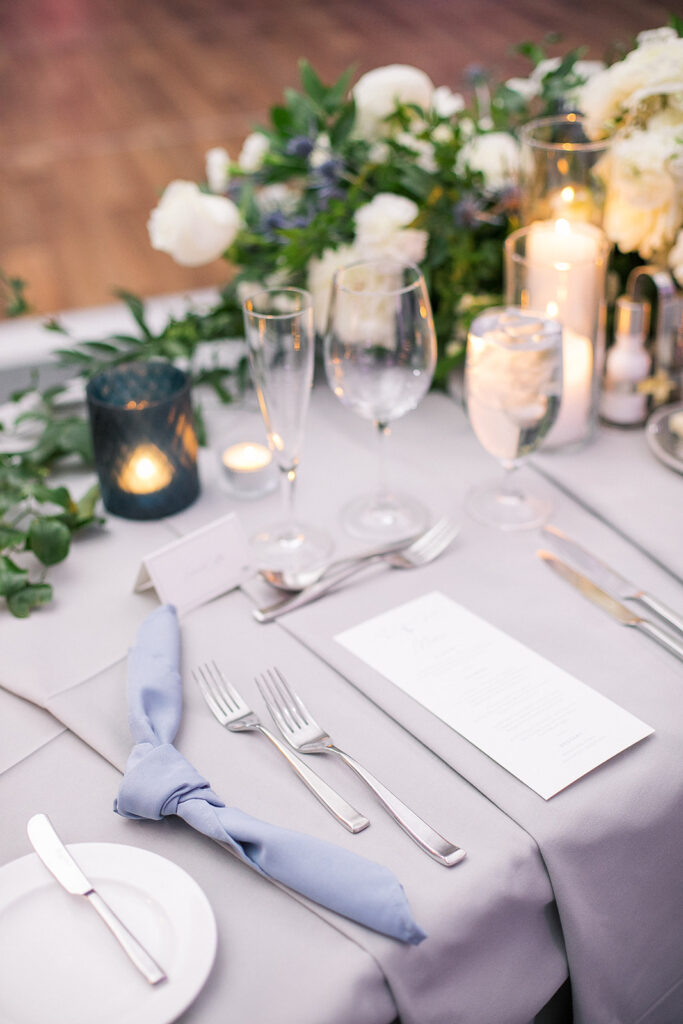 Reception guest place setting with gray a d blue linens, candles, and white floral and greenery centerpieces.