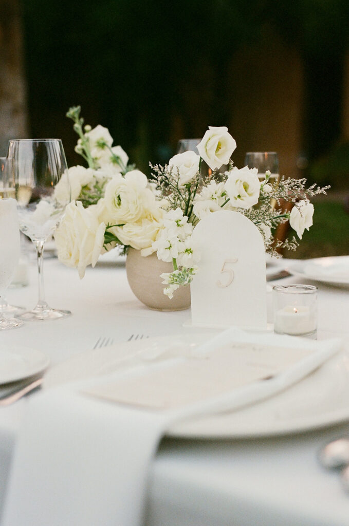 White linens and white floral arrangements with table number and votive candle on reception table.
