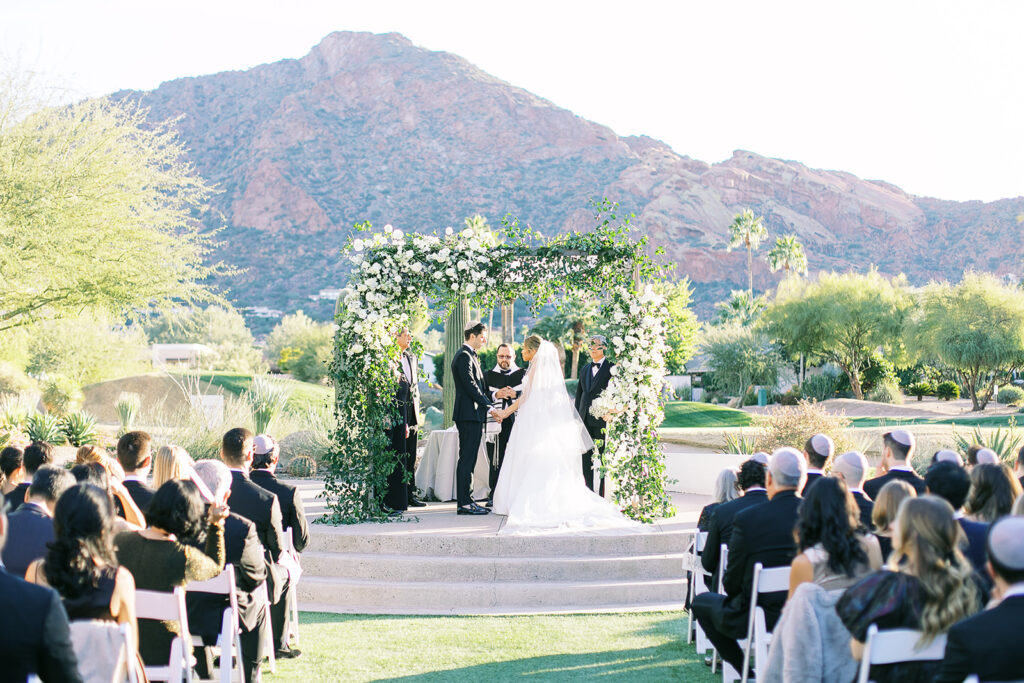 Bride and groom holding hands, standing under chuppah with with flowers and greenery at Mountain Shadows at outdoor ceremony.