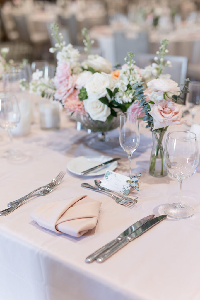 Wedding reception table with glass vessel centerpiece of white and blush roses and bud vases on blush linens.
