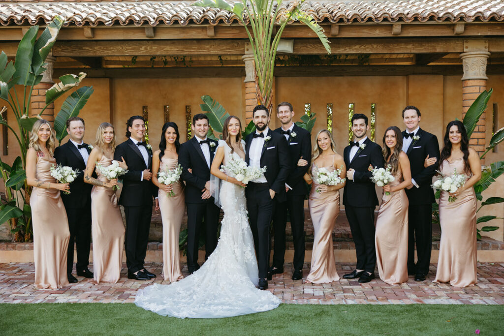 Wedding party standing on bricked walkway at Royal Palms with bridesmaids wearing rose gold gowns and groomsmen in black, alternating where they are standihng and bride and groom in center.