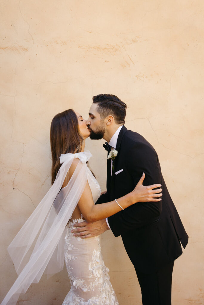 Bride and groom kissing in front of smooth stucco wall.