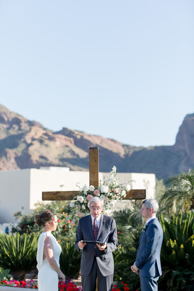 Outdoor wedding ceremony with desert mountains in the background and bride and groom with officiant standing in front of wooden cross with pink and blush floral installation.