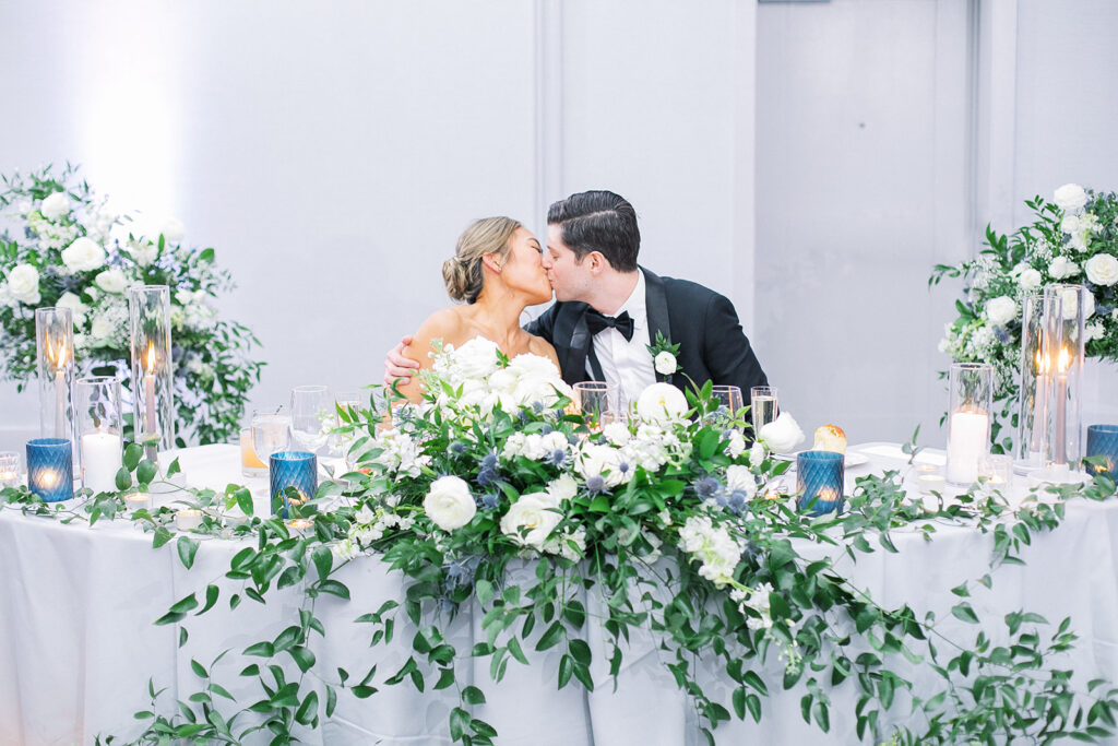 Bride and groom kissing while sitting at sweetheart table at indoor wedding reception.