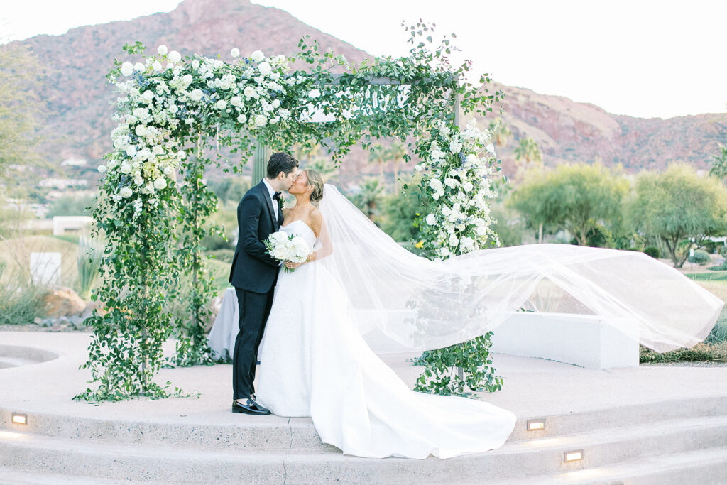 Bride and groom kissing in front of wedding ceremony altar space with bride's veil blowing out to side behind her.