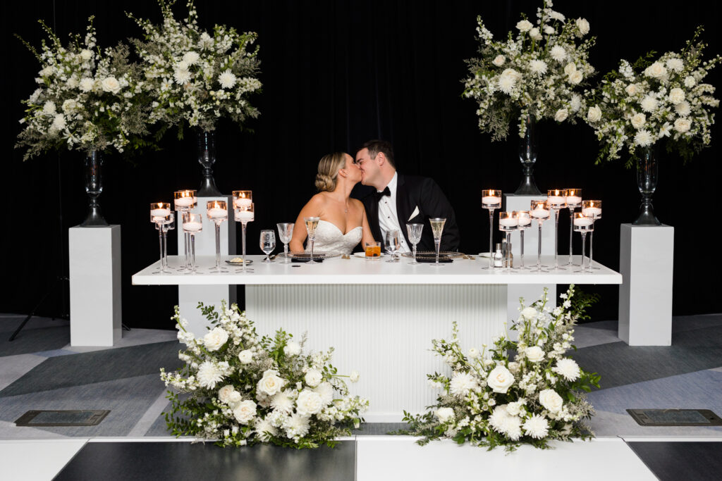 Bride and groom kissing, seated at wedding reception sweetheart table with candles and floral framing them in back and on ground in front of table.