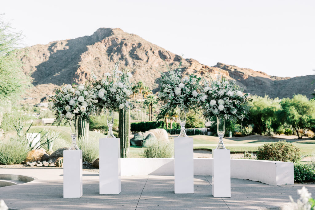 Four floral arrangements for outdoor wedding ceremony altar space in tall glass vases resting on two different size white acrylic pillars.