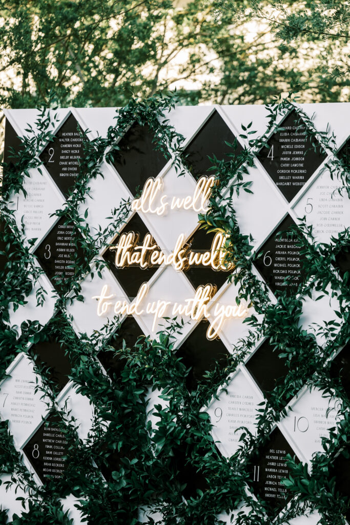 Black and white diamond board with greenery escort board with neon sign in middle.