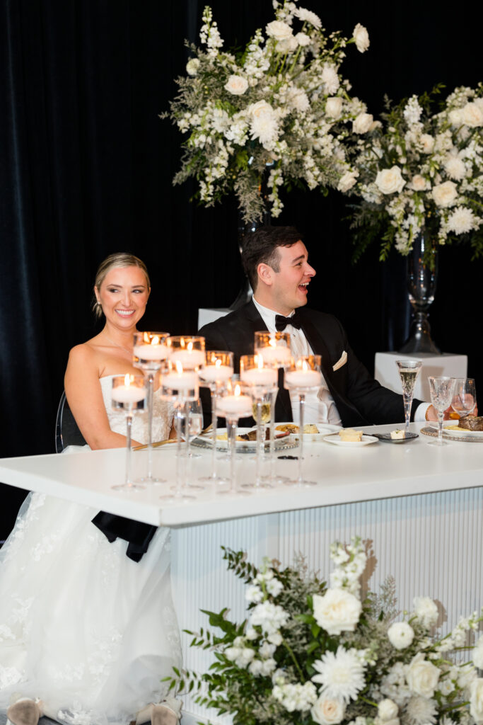 Bride and groom smiling, sitting at sweetheart table with candles and white floral arrangements.